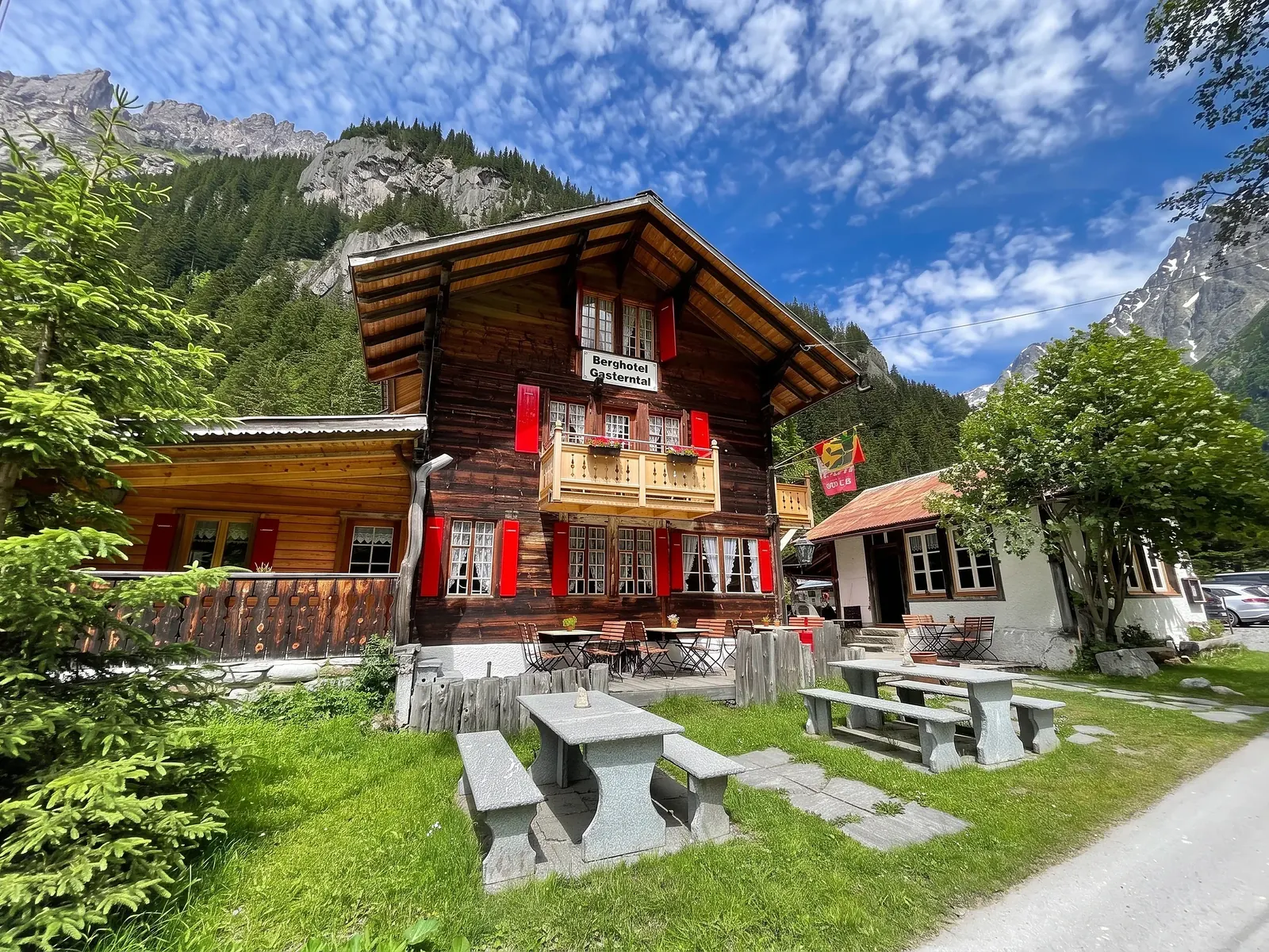 Giebelseite mit Balkon, Geranien und Hausschild, davor Steintische